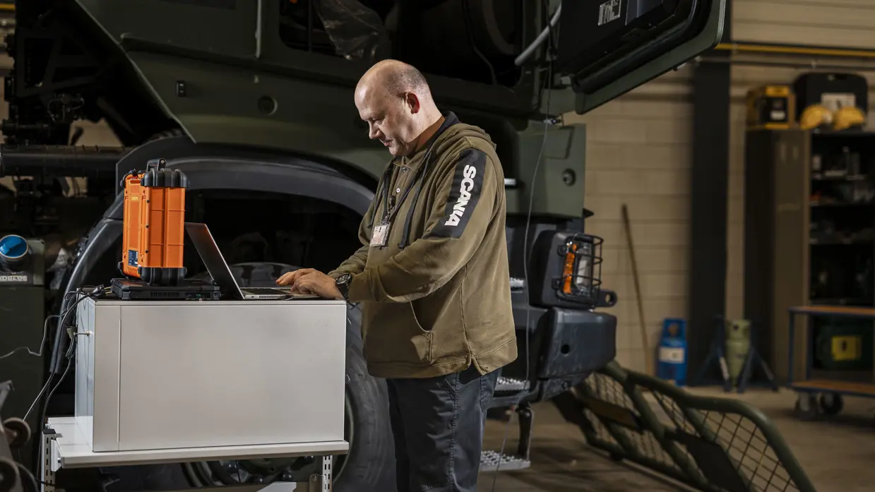 Man kijkt op laptop en staat naast transportvoertuig in werkplaats.