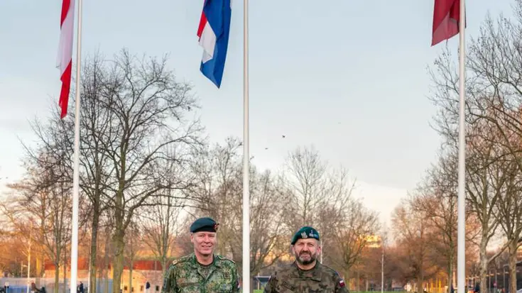 2 militairen op een grasveld met 3 vlaggen op de achtergrond.