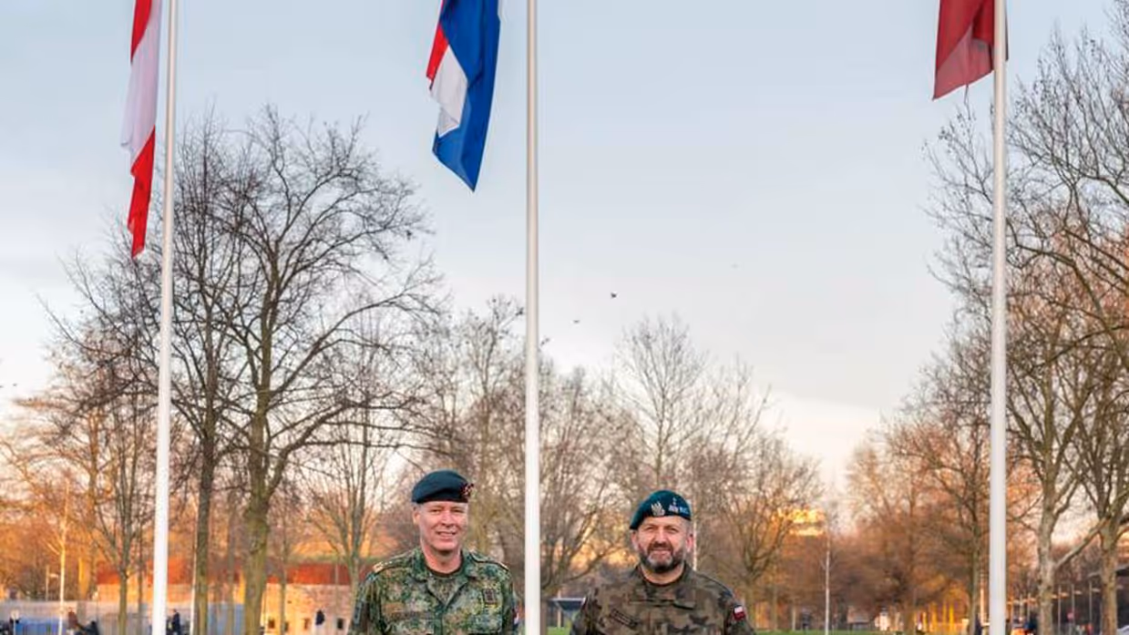 2 militairen op een grasveld met 3 vlaggen op de achtergrond.