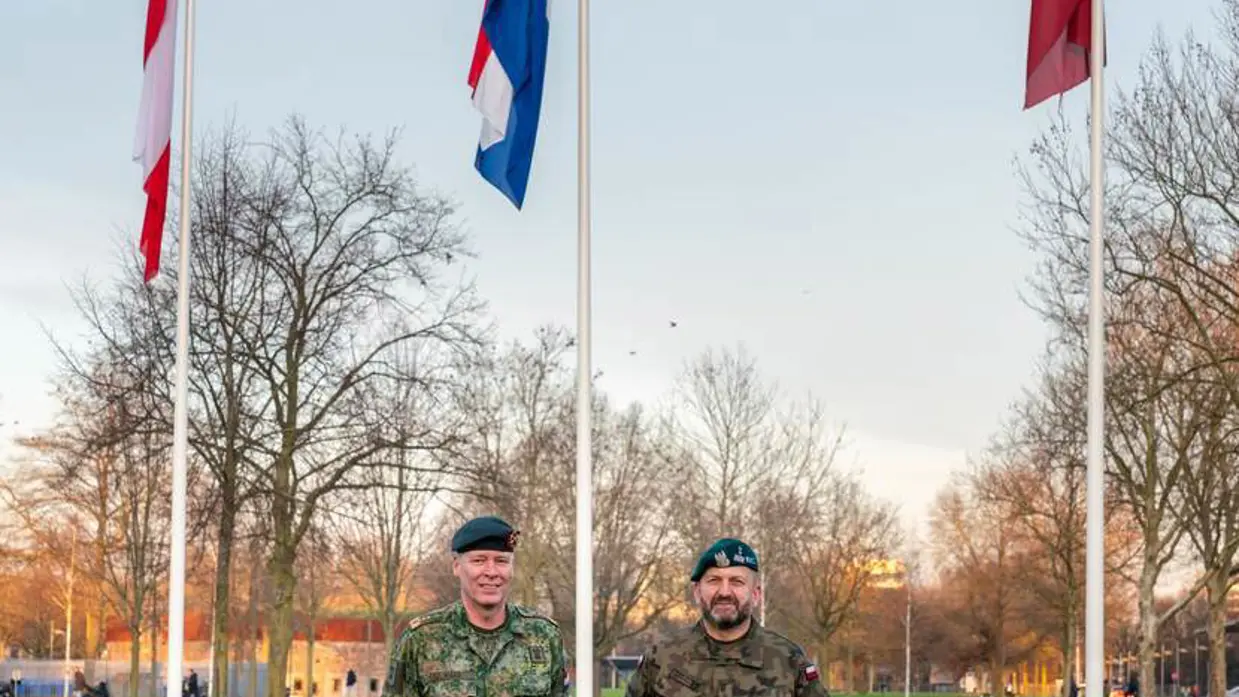 2 militairen op een grasveld met 3 vlaggen op de achtergrond.