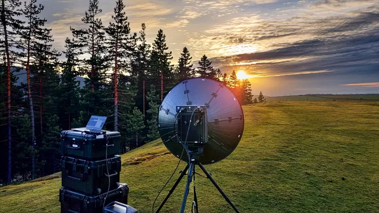 Een schotelantenne in een weiland naast bomen met op de achtergrond een ondergaande zon.