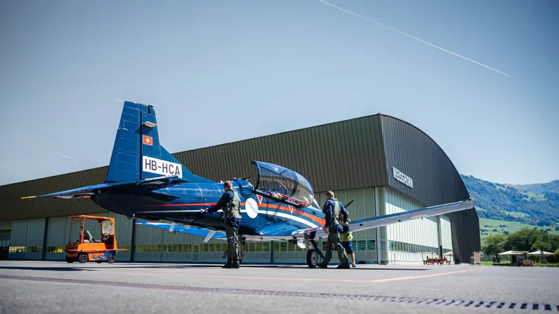 Twee vliegers verrichten handelingen aan het vliegtuig dat voor een grote grijze hangar met een rond dak staat.