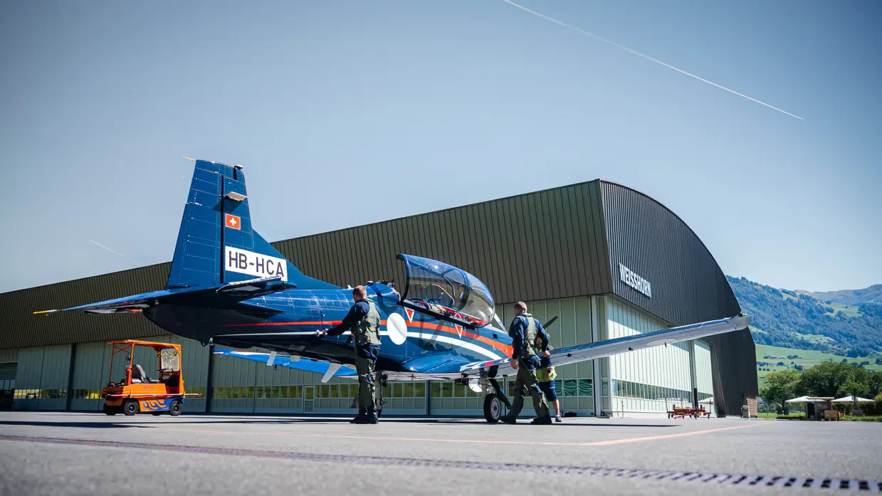 Twee vliegers verrichten handelingen aan het vliegtuig dat voor een grote grijze hangar met een rond dak staat.