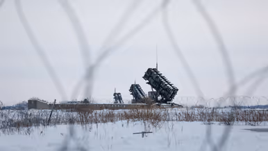 Op de linkerfoto opent luitenant Leon een deur van een groene werkcontainer. Op de rechterfoto is een Patriot-lanceerinrichting te zien door het prikkeldraad heen.