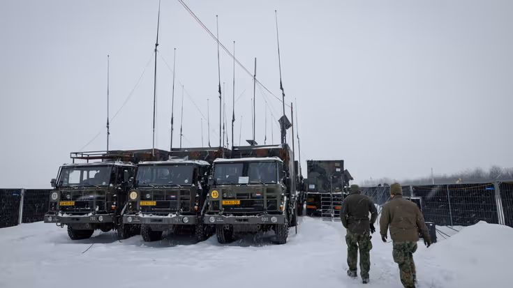 Twee militairen lopen door een sneeuwbui richting enkele groene vrachtwagens met veel antennes.
