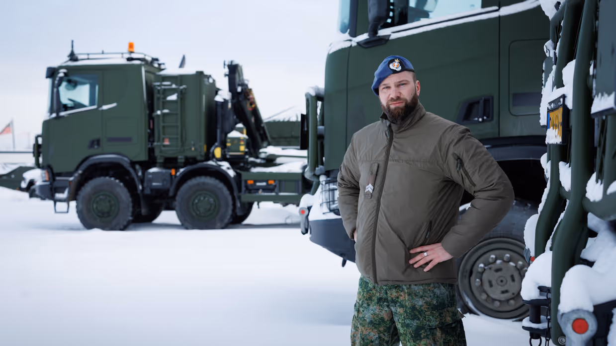 Portretfoto van Rick met zijn handen in zijn zij. Op de achtergrond enkele militaire vrachtwagens.