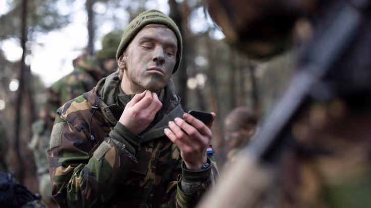 Een deelnemer brengt camouflagekleuren op op zijn gezicht. Hij heeft 1 hand bij zijn kin.