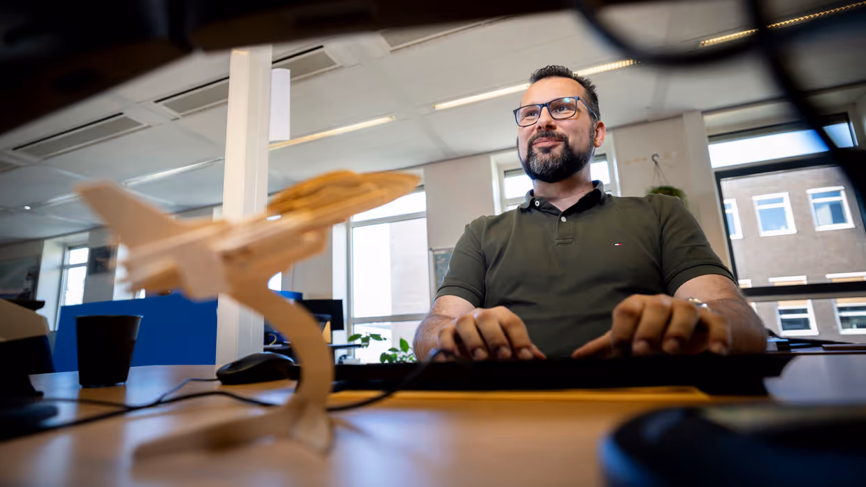 Marcel achter zijn bureau, kijkend naar zijn beeldscherm met op de voorgrond een houten vliegtuig.