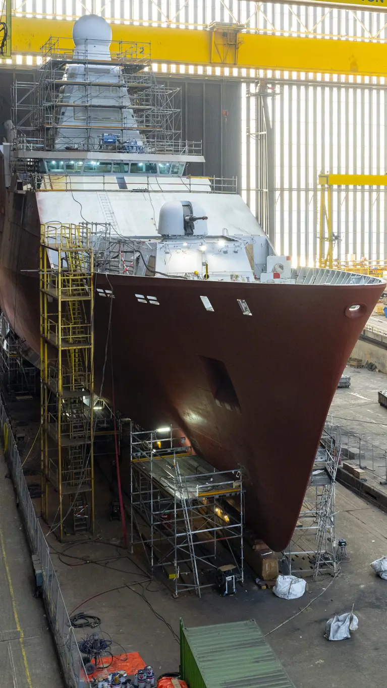 Zr.Ms. Holland in aanbouw in een overdekte scheepswerf, omringd door steigers, kranen en bouwmateriaal.