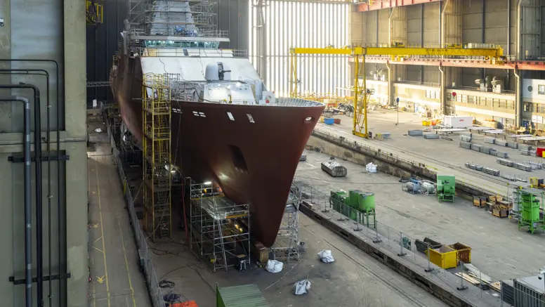 Zr.Ms. Holland in aanbouw in een overdekte scheepswerf, omringd door steigers, kranen en bouwmateriaal.
