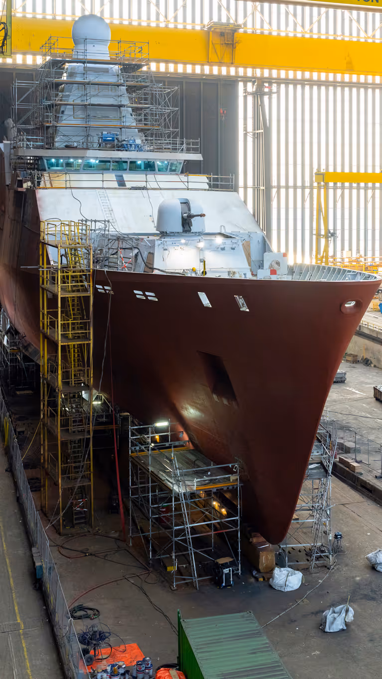 Zr.Ms. Holland in aanbouw in een overdekte scheepswerf, omringd door steigers, kranen en bouwmateriaal.