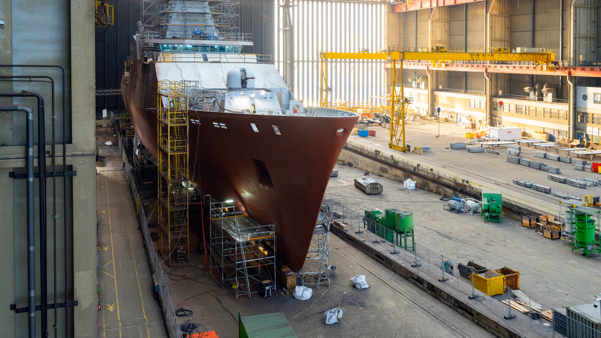Zr.Ms. Holland in aanbouw in een overdekte scheepswerf, omringd door steigers, kranen en bouwmateriaal.
