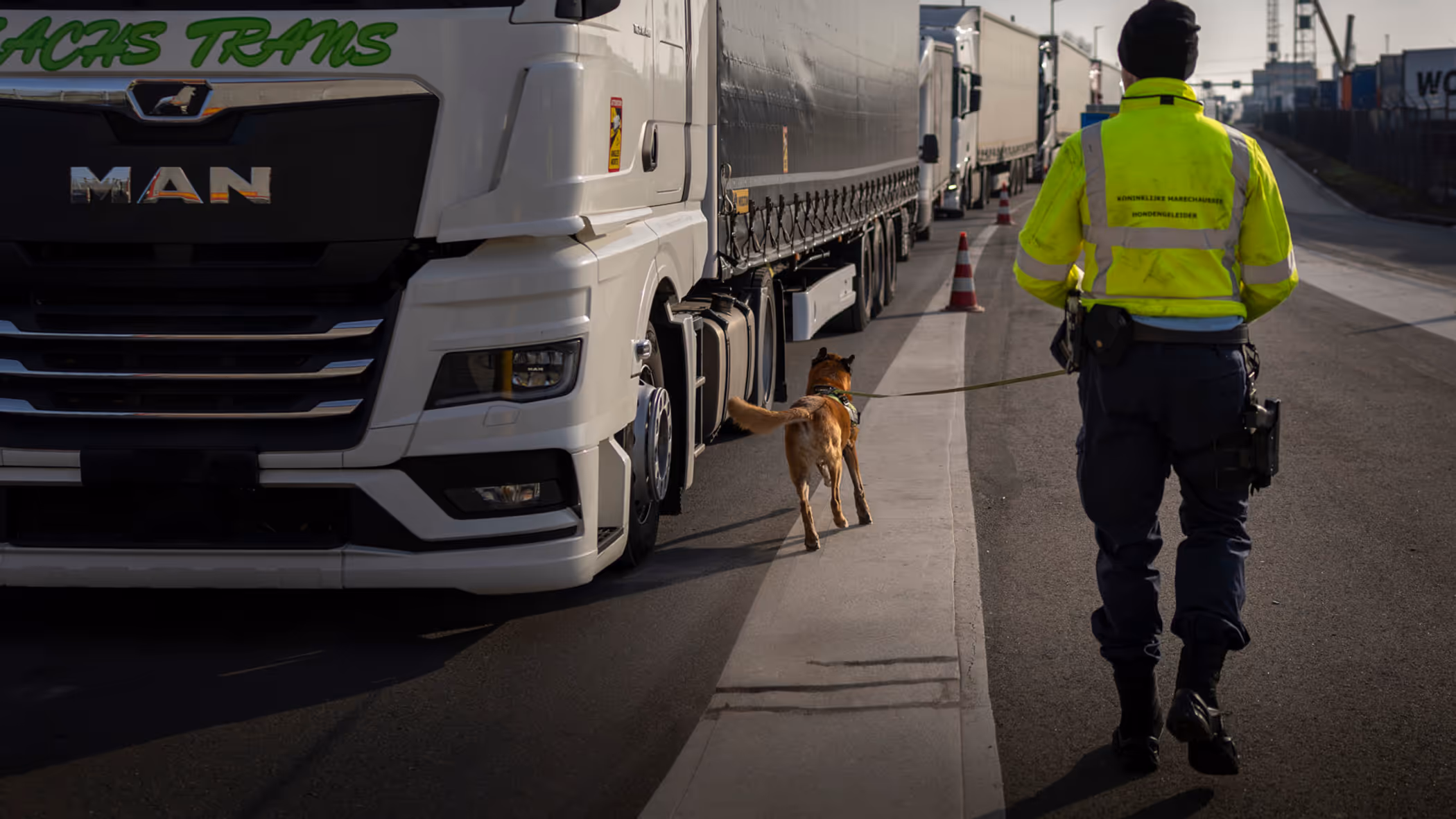 Marechaussee met hond op de rug gezien loopt langs trailer.