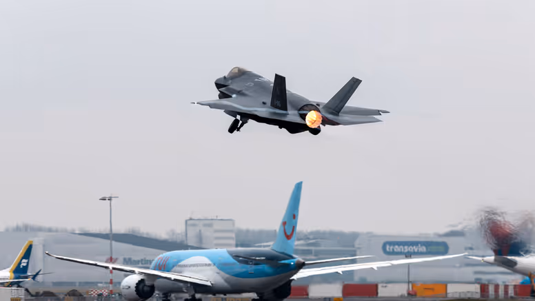 Een F-35 gevechtsvliegtuig stijgt op vanaf luchthaven Schiphol. Op de achtergrond is de luchthaven en een taxiënd civiel passagiersvliegtuig te zien.