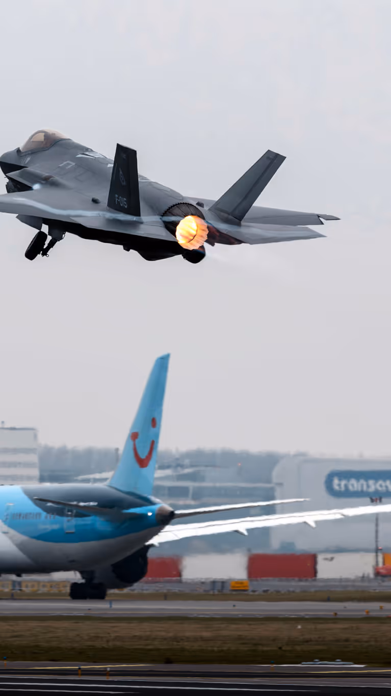 Een F-35 gevechtsvliegtuig stijgt op vanaf luchthaven Schiphol. Op de achtergrond is de luchthaven en een taxiënd civiel passagiersvliegtuig te zien.