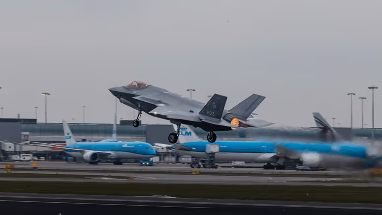 Een opstijgende F-35 op luchthaven Schiphol. Op de achtergrond zijn geparkeerde blauwe toestellen van KLM te zien.&nbsp;
