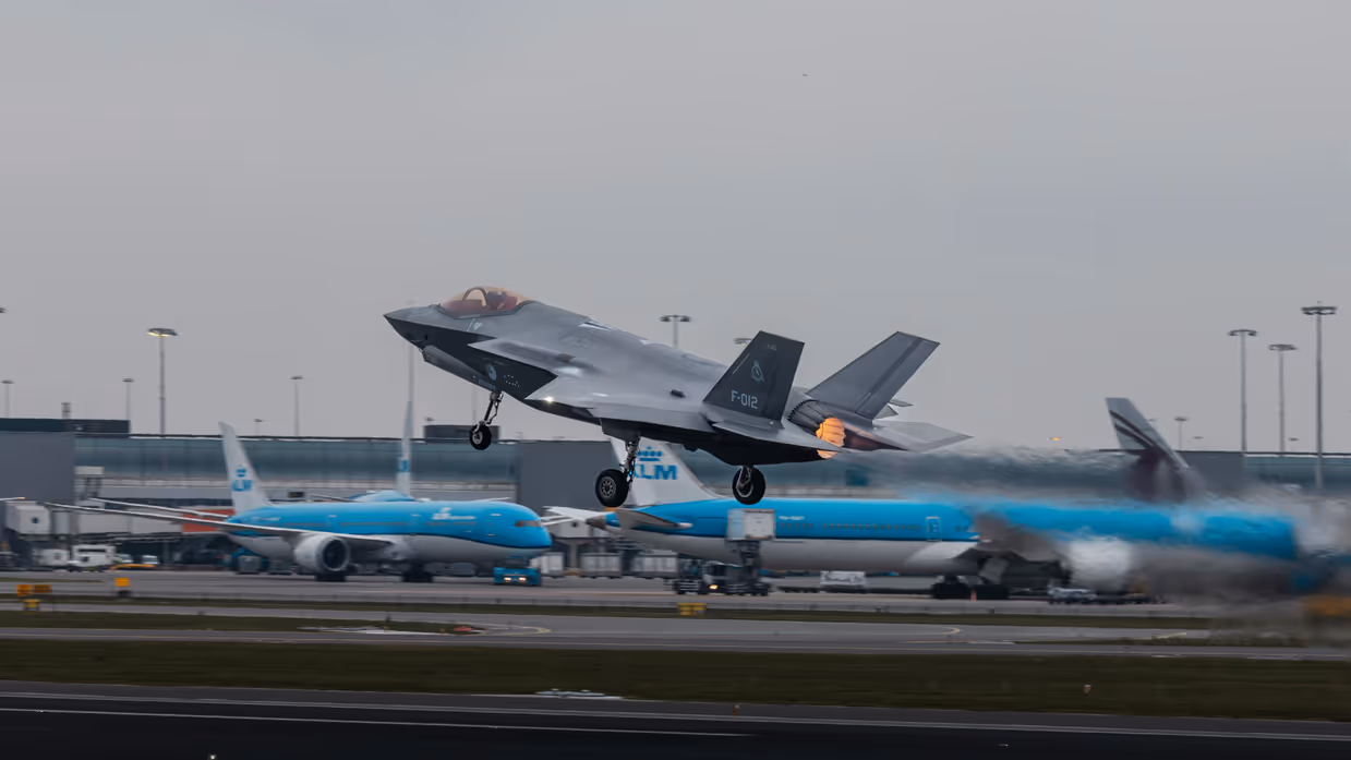 Een opstijgende F-35 op luchthaven Schiphol. Op de achtergrond zijn geparkeerde blauwe toestellen van KLM te zien.&nbsp;