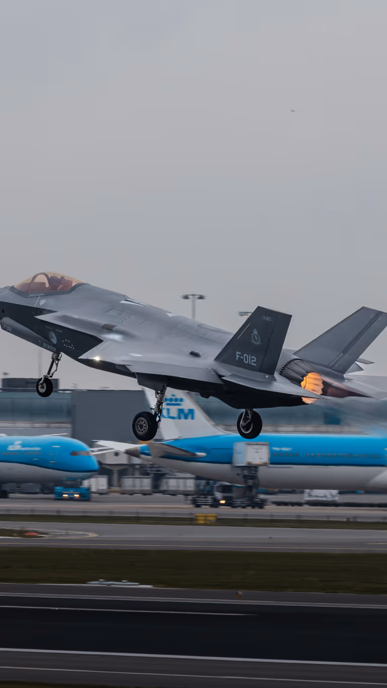 Een opstijgende F-35 op luchthaven Schiphol. Op de achtergrond zijn geparkeerde blauwe toestellen van KLM te zien.&nbsp;