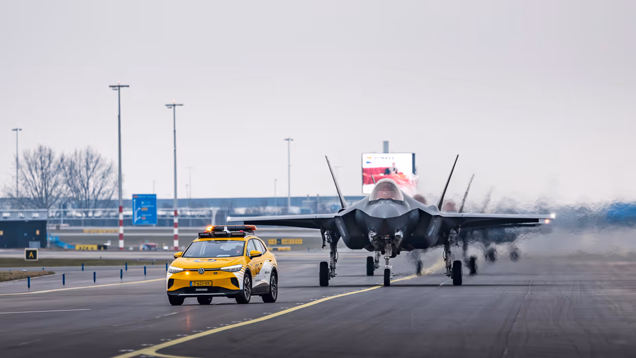 Een geparkeerde F-35 staat op luchthaven Schiphol, met de klep van de cockpit geopend. Om het toestel bewegen militairen, die onderhoud plegen. Op de voorgrond loopt iemand met een schijf met de tekst ‘remove before flight’. De luchtmachters dragen een groene militaire broek, met daarboven een oranje veiligheidshesje. Daarnaast staat een foto van taxiende F-35’s op het vliegveld. Op de achtergrond is de snelweg te zien.