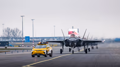 Een geparkeerde F-35 staat op luchthaven Schiphol, met de klep van de cockpit geopend. Om het toestel bewegen militairen, die onderhoud plegen. Op de voorgrond loopt iemand met een schijf met de tekst ‘remove before flight’. De luchtmachters dragen een groene militaire broek, met daarboven een oranje veiligheidshesje. Daarnaast staat een foto van taxiende F-35’s op het vliegveld. Op de achtergrond is de snelweg te zien.