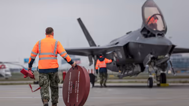 Een geparkeerde F-35 staat op luchthaven Schiphol, met de klep van de cockpit geopend. Om het toestel bewegen militairen, die onderhoud plegen. Op de voorgrond loopt iemand met een schijf met de tekst ‘remove before flight’. De luchtmachters dragen een groene militaire broek, met daarboven een oranje veiligheidshesje. Daarnaast staat een foto van taxiende F-35’s op het vliegveld. Op de achtergrond is de snelweg te zien.