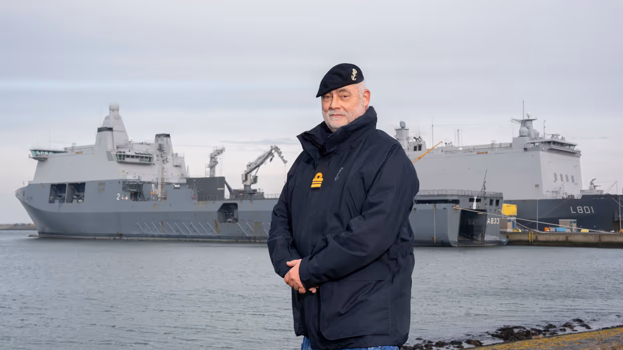 Alttekst: Een marineman poseert aan de kade voor 2 marineschepen. 