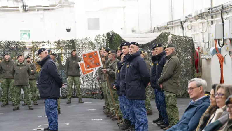 Militairen van de landmacht en marine staan naast elkaar gepositioneerd in een hal, met op de achtergrond enkele burgers. 