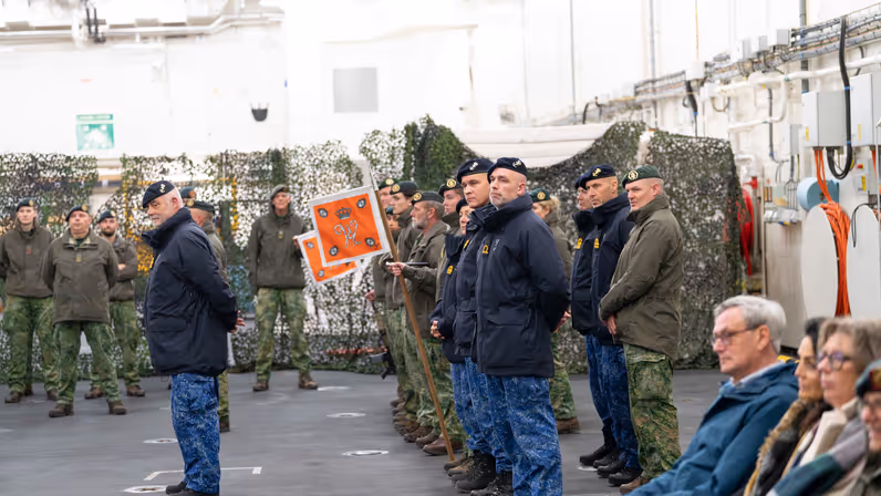 Militairen van de landmacht en marine staan naast elkaar gepositioneerd in een hal, met op de achtergrond enkele burgers. 