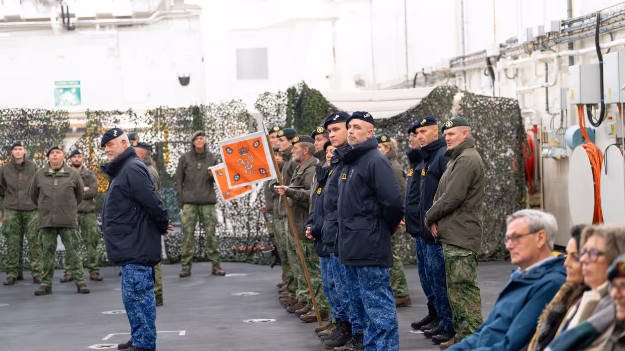 Militairen van de landmacht en marine staan naast elkaar gepositioneerd in een hal, met op de achtergrond enkele burgers. 