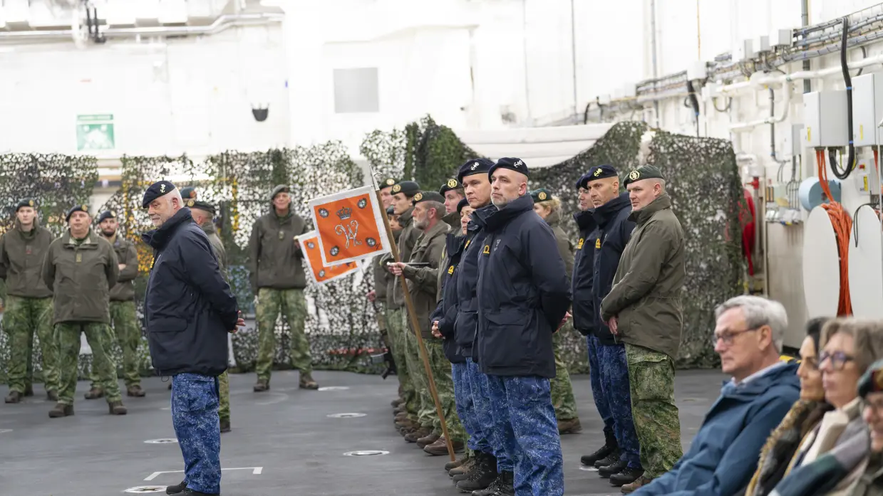 Militairen van de landmacht en marine staan naast elkaar gepositioneerd in een hal, met op de achtergrond enkele burgers. 