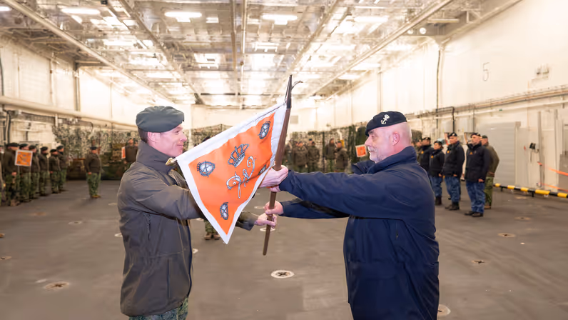Een militair van de marine krijgt een vlag overhandigd uit handen van een militair van de landmacht. 