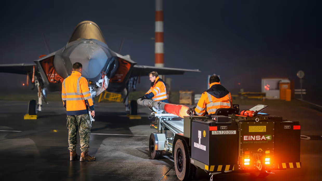 Militairen bewegen met oefenbewapening op een kar naar een geparkeerde F-35. Het is donker, wat een spannend schouwspel creëert.