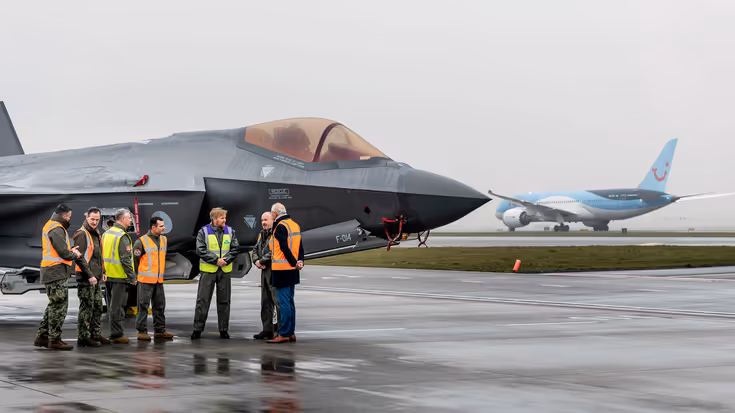 Een groep mensen staat met elkaar te praten voor een geparkeerd gevechtsvliegtuig. In het midden van het gezelschap staat koning Willem Alexander, die een geel veiligheidshesje draagt op zijn groene vliegerspak. In de achtergrond is een geparkeerd passagierstoestel van KLM te zien.