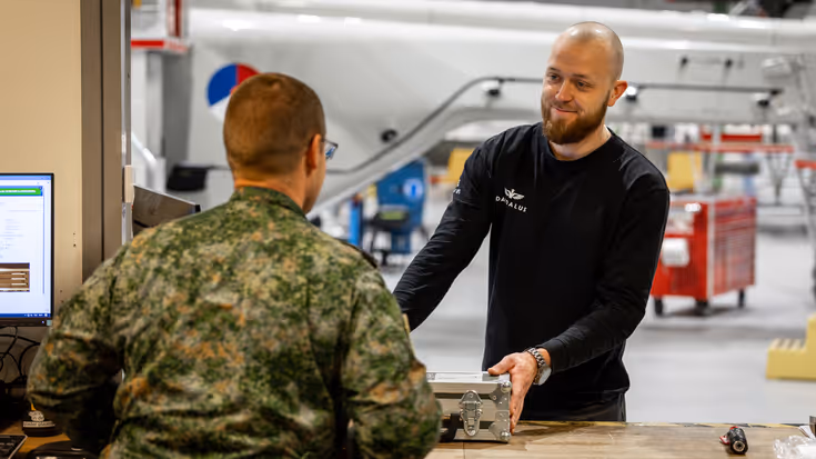 Aan de balie geeft een militaire medewerker een koffertje met apparatuur af aan een burger collega.