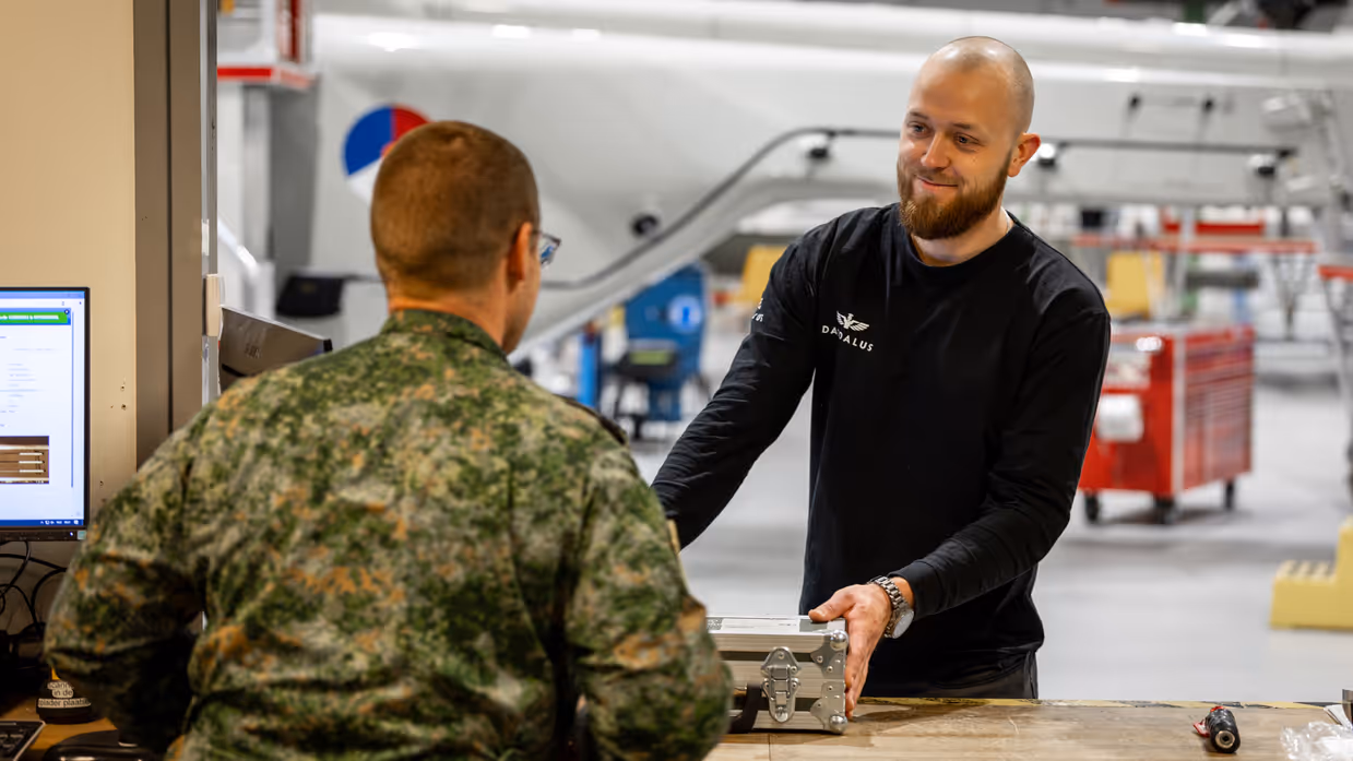 Aan de balie geeft een militaire medewerker een koffertje met apparatuur af aan een burger collega.
