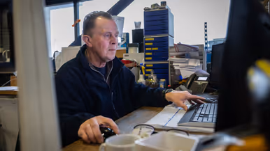 Een medewerker monitort van dichtbij hoe een pallet wordt uitgeladen. Op de tweede foto een medewerker, gezeten achter zijn bureau.