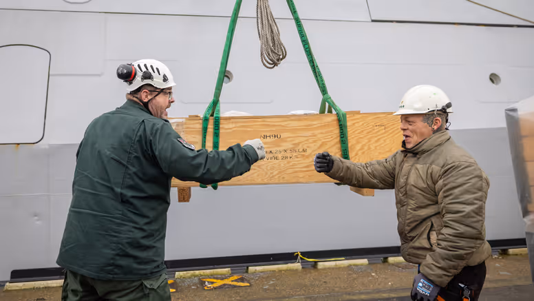 Twee collega’s geven elkaar een boks, met op de achtergrond een houten box, waarop NH90 staat.