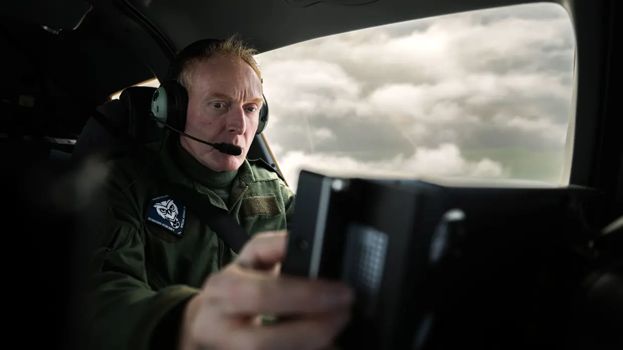 Een piloot zit in de cockpit van het patrouillevliegtuig achter het stuur, rechts van hem is een raam met uitzicht op het wolkendek.