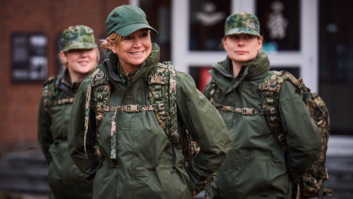 Een lachende koningin Máxima in gevechtstenue met pet op en rugzak om, achter haar twee vrouwelijke militairen in dezelfde outfit.