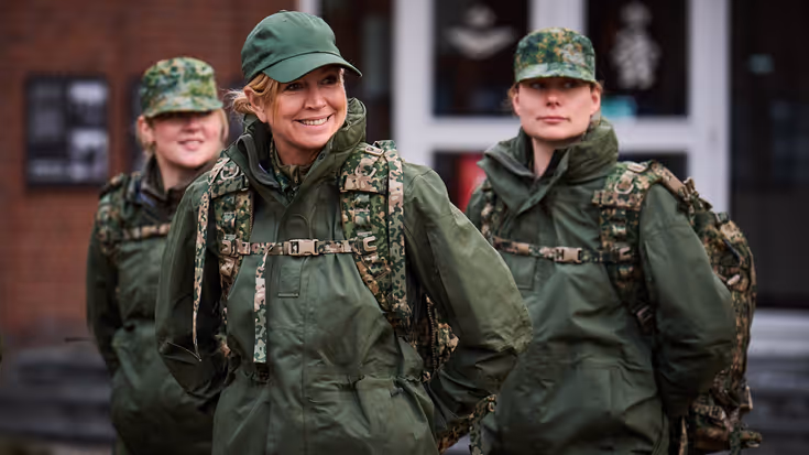 Een lachende koningin Máxima in gevechtstenue met pet op en rugzak om, achter haar twee vrouwelijke militairen in dezelfde outfit.