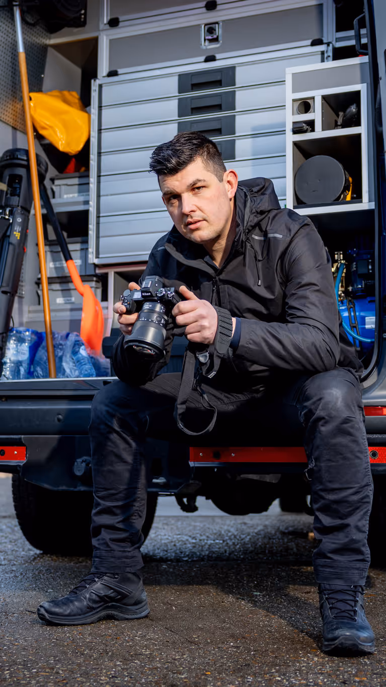 Adjudant Allard zit achterin de dienstbus met zijn camera vast en een blik naar de camera toe.