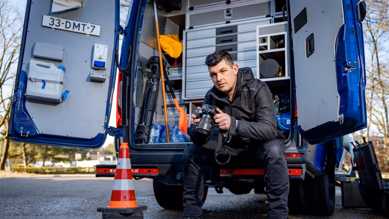 Adjudant Allard zit achterin de dienstbus met zijn camera vast en een blik naar de camera toe.