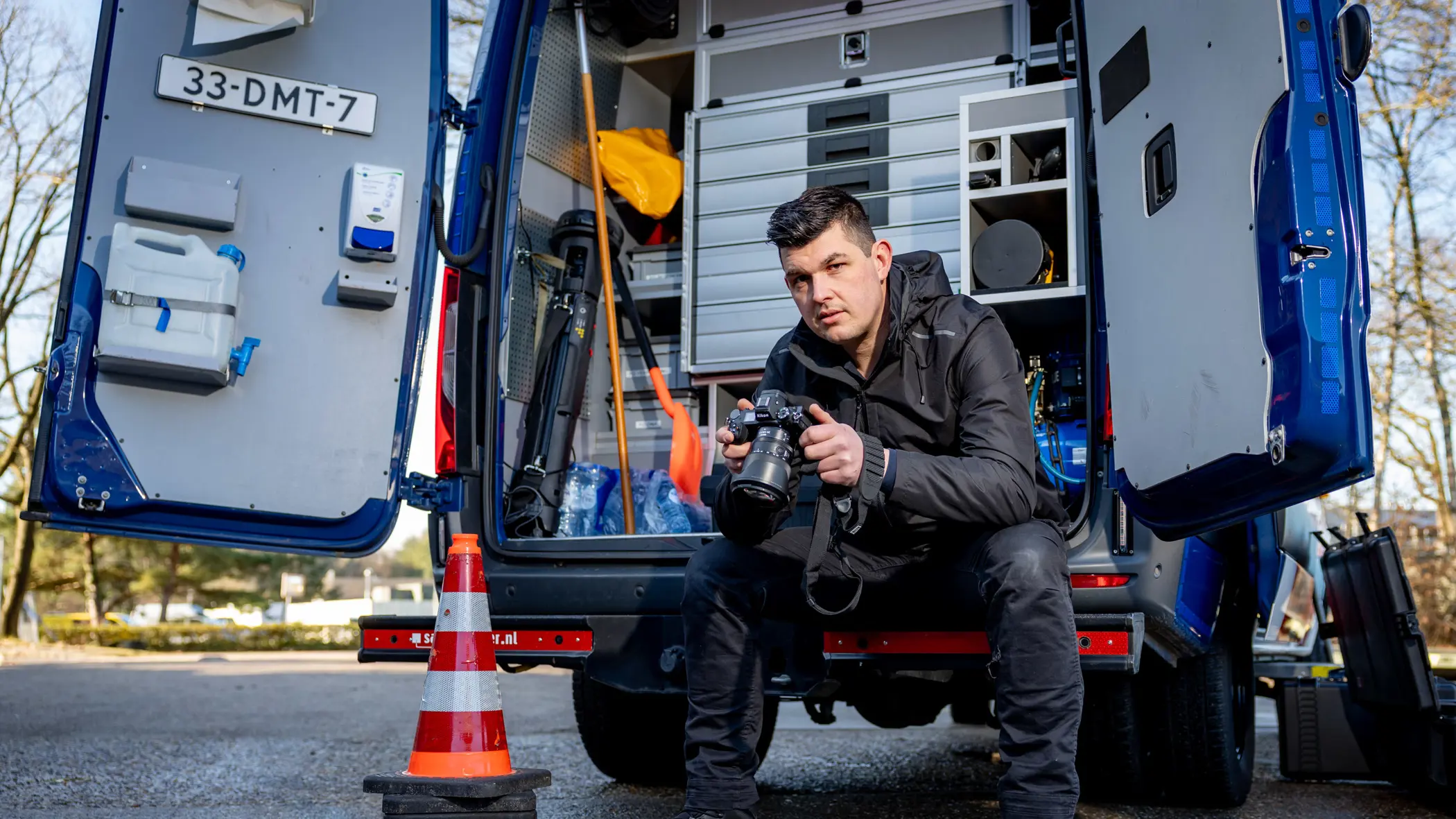 Adjudant Allard zit achterin de dienstbus met zijn camera vast en een blik naar de camera toe.