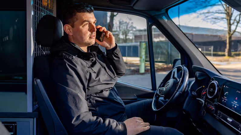 Adjudant Allard op de bestuurdersstoel van de dienstbus, met een mobiele telefoon aan zijn oor.