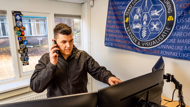 Adjudant Allard staat in zijn nieuwe kantoor en wijst naar een van zijn schermen terwijl hij aan het bellen is.