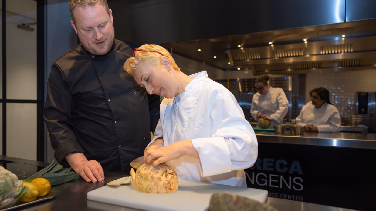 Dijan staat in een keuken en snijdt onder toeziend oog van een docent een knol. 