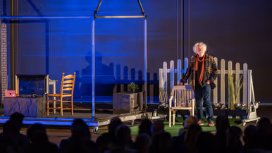 Op een theatervloer staat een stalen geraamte van een huis met daarin een nagebootste woonkamer. Ernaast een tuin waarin een man naast een tuinstoel staat, hij kijkt omhoog.