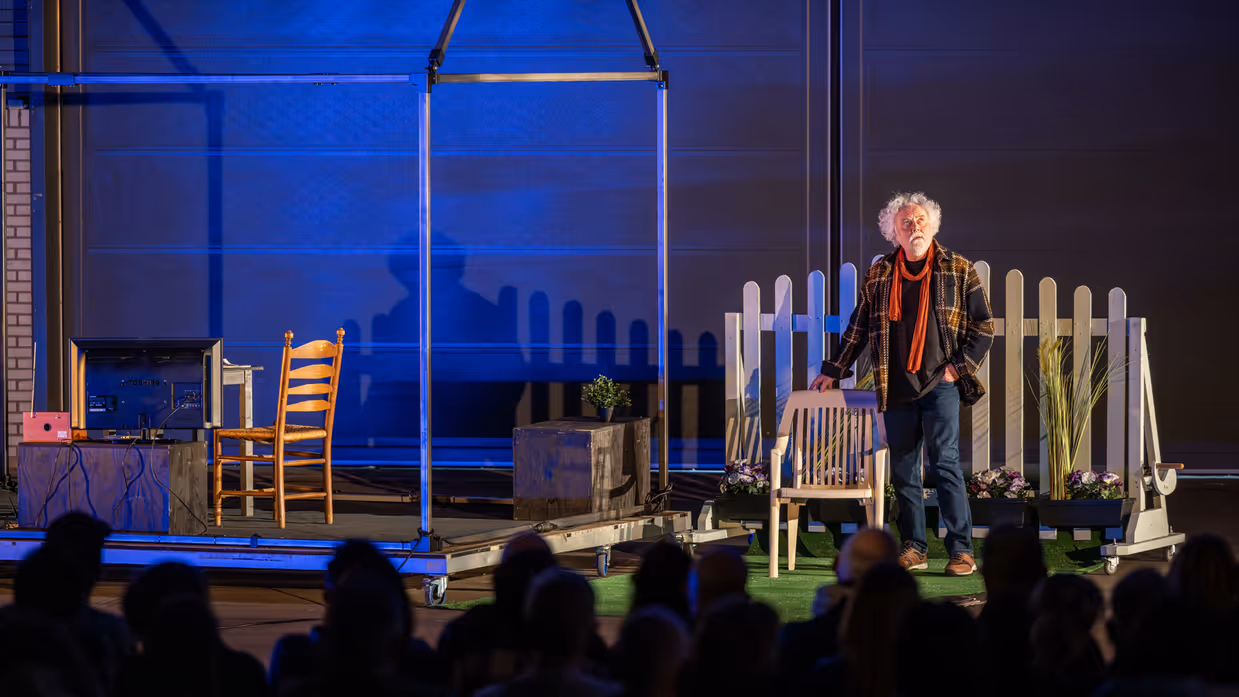Op een theatervloer staat een stalen geraamte van een huis met daarin een nagebootste woonkamer. Ernaast een tuin waarin een man naast een tuinstoel staat, hij kijkt omhoog.