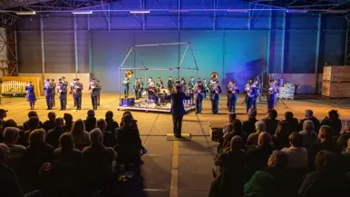 In een hangaar zitten een aantal rijen mensen gescheiden door een gangpad. Ze kijken naar een orkest met luchtmachtmilitairen met daarvoor een dirigent. Het orkest staat opgesteld rondom een stalen geraamte van een huis waarin een man zit.