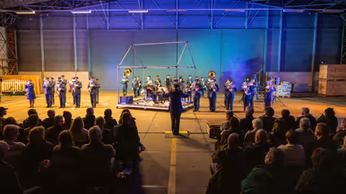 In een hangaar zitten een aantal rijen mensen gescheiden door een gangpad. Ze kijken naar een orkest met luchtmachtmilitairen met daarvoor een dirigent. Het orkest staat opgesteld rondom een stalen geraamte van een huis waarin een man zit.